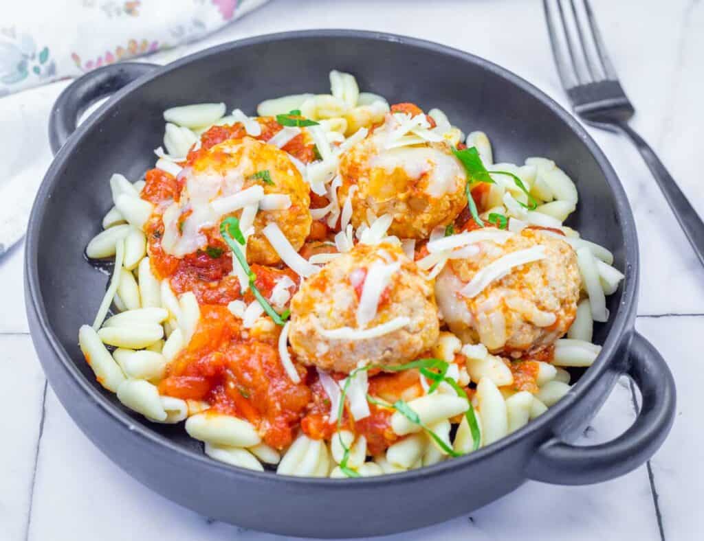 A black bowl filled with pasta, topped with tomato sauce, meatballs, shredded cheese, and garnished with fresh herbs. A fork and napkin are placed nearby.