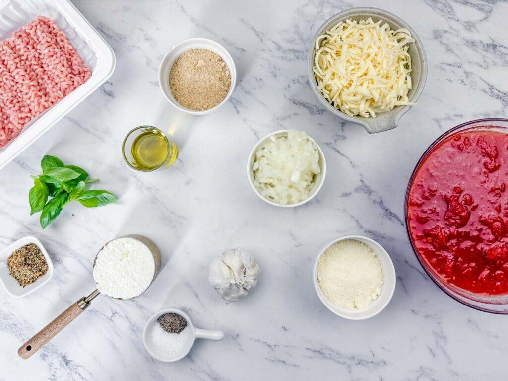 Various ingredients for cooking, including ground meat, shredded cheese, diced onions, tomato sauce, grated cheese, garlic, oil, breadcrumbs, basil, flour, and seasonings on a marble surface.