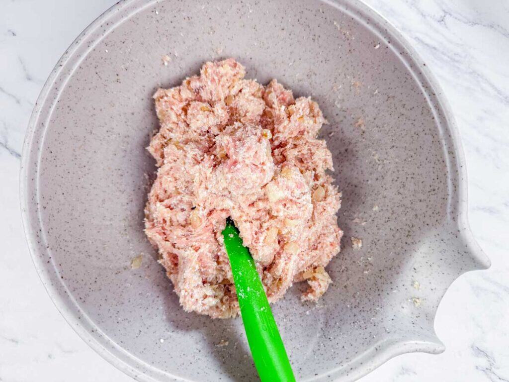 A mixing bowl containing raw ground meat and seasonings being stirred with a green spatula.