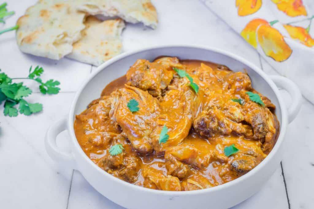 A white bowl filled with chicken curry garnished with cilantro, with pieces of flatbread and cilantro on the side.