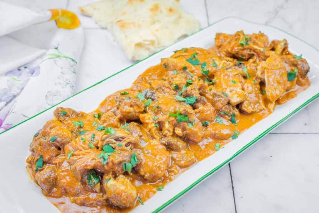 A rectangular white plate filled with chicken pieces in orange curry sauce, garnished with chopped herbs, with naan bread in the background.