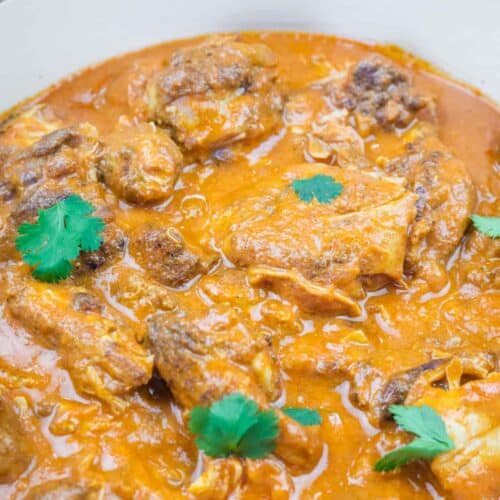 A pot of chicken pieces cooked in a thick, orange-red curry sauce, garnished with fresh cilantro leaves.