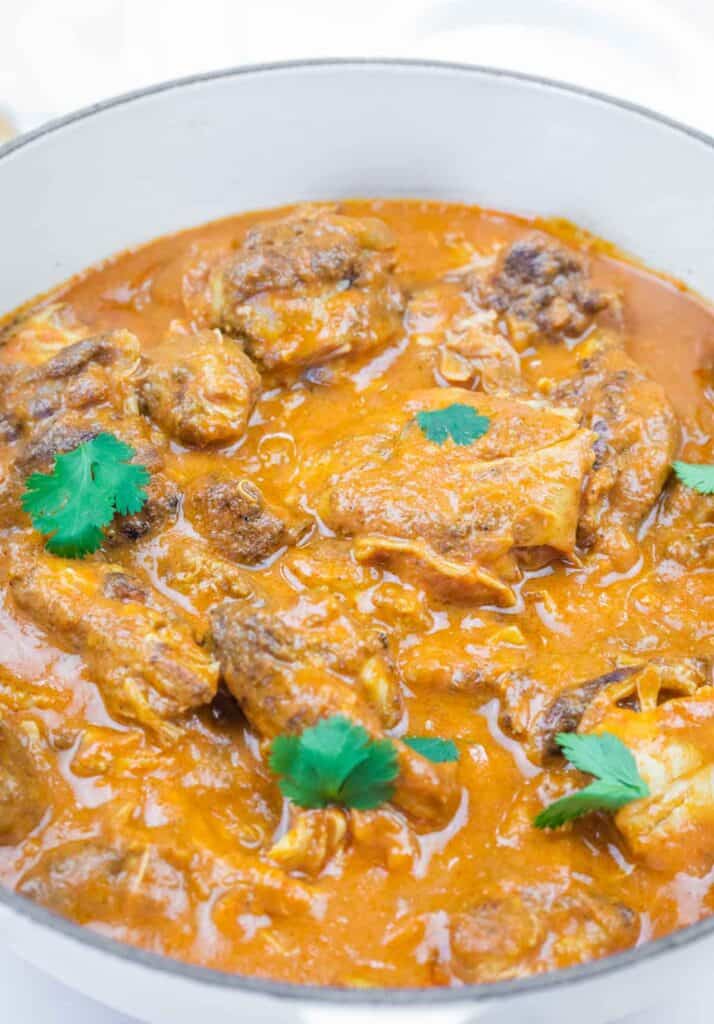 A pot of chicken pieces cooked in a thick, orange-red curry sauce, garnished with fresh cilantro leaves.
