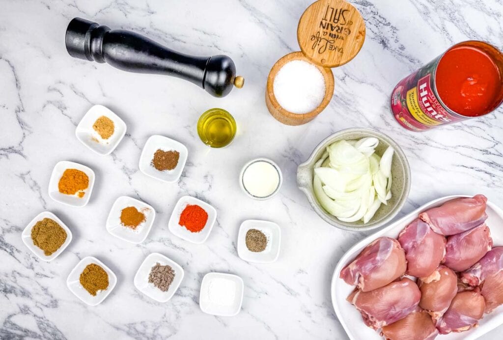 Chicken thighs, sliced onions, canned tomatoes, various spices in small bowls, oil, cream, salt, and a pepper mill arranged on a marble countertop.