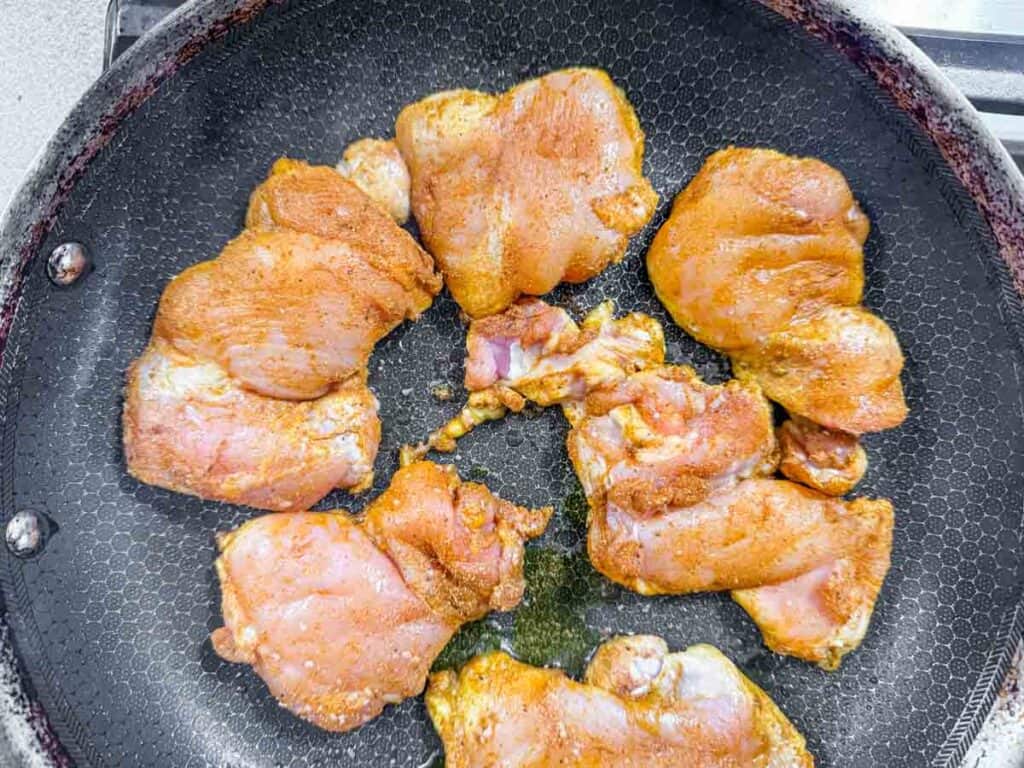 Raw, seasoned chicken thighs arranged in a single layer on a nonstick frying pan, ready to be cooked.