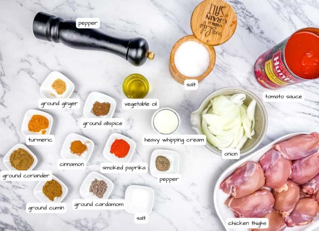 Various labeled ingredients for a recipe are arranged on a marble surface, including spices, chicken thighs, onion, tomato sauce, oil, cream, salt, and pepper.