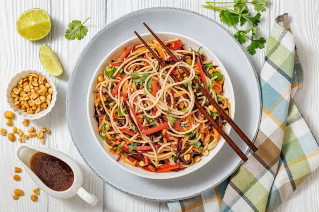 A bowl of noodle salad with vegetables, garnished with herbs, is placed on a plate with chopsticks, alongside peanuts, lime wedges, sauce, and a checkered napkin.