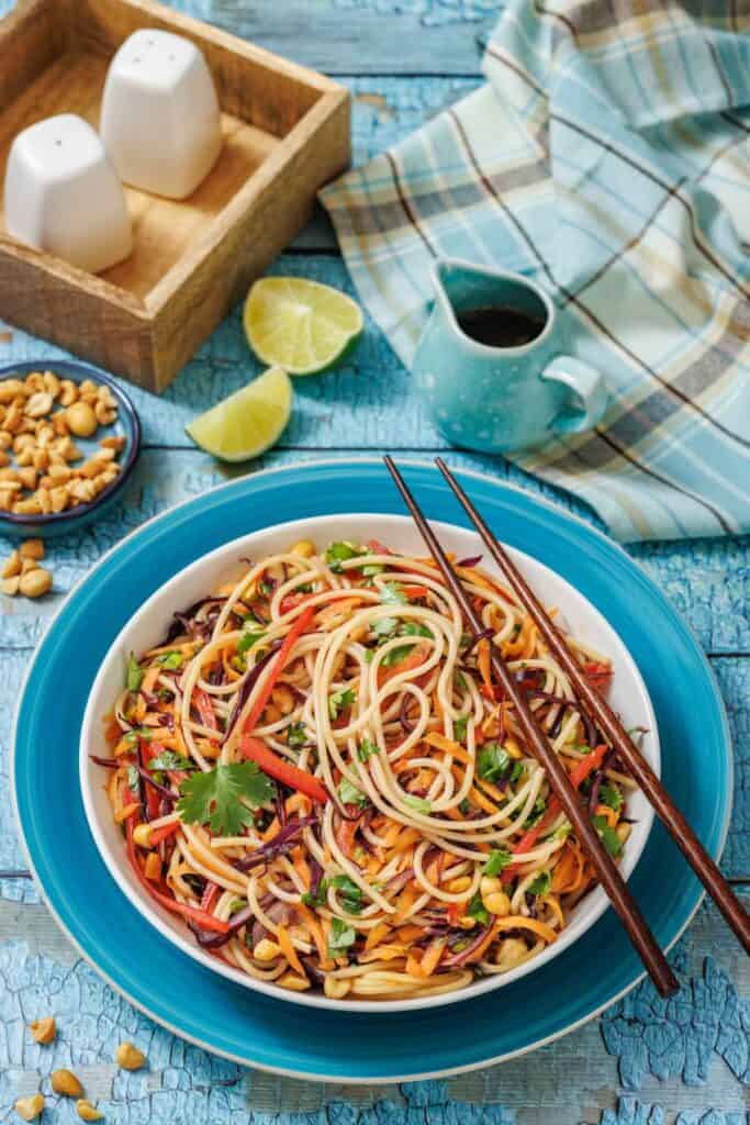 A bowl of colorful noodle salad with vegetables and chopsticks, served on a blue plate, with peanuts, lime wedges, and sauce on a rustic blue table.