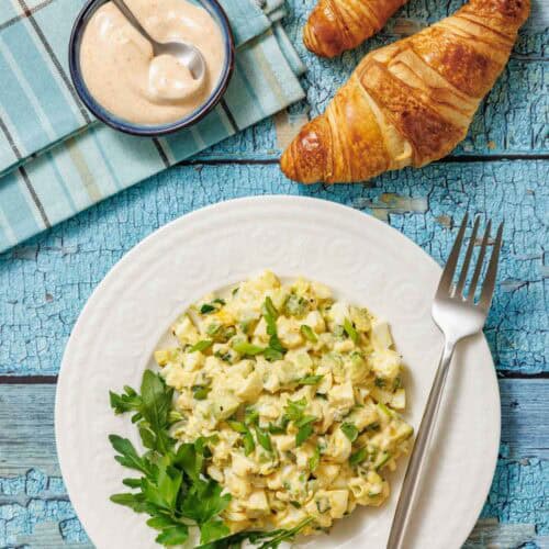 A white plate with avocado egg salad, garnished with greens, is shown with croissants, a bowl of sauce, and a cloth on a blue wooden surface.