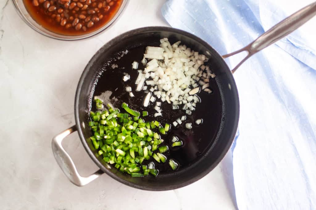 A black saucepan contains chopped onions and green peppers in oil, with a bowl of baked beans and a blue cloth nearby on a light countertop.