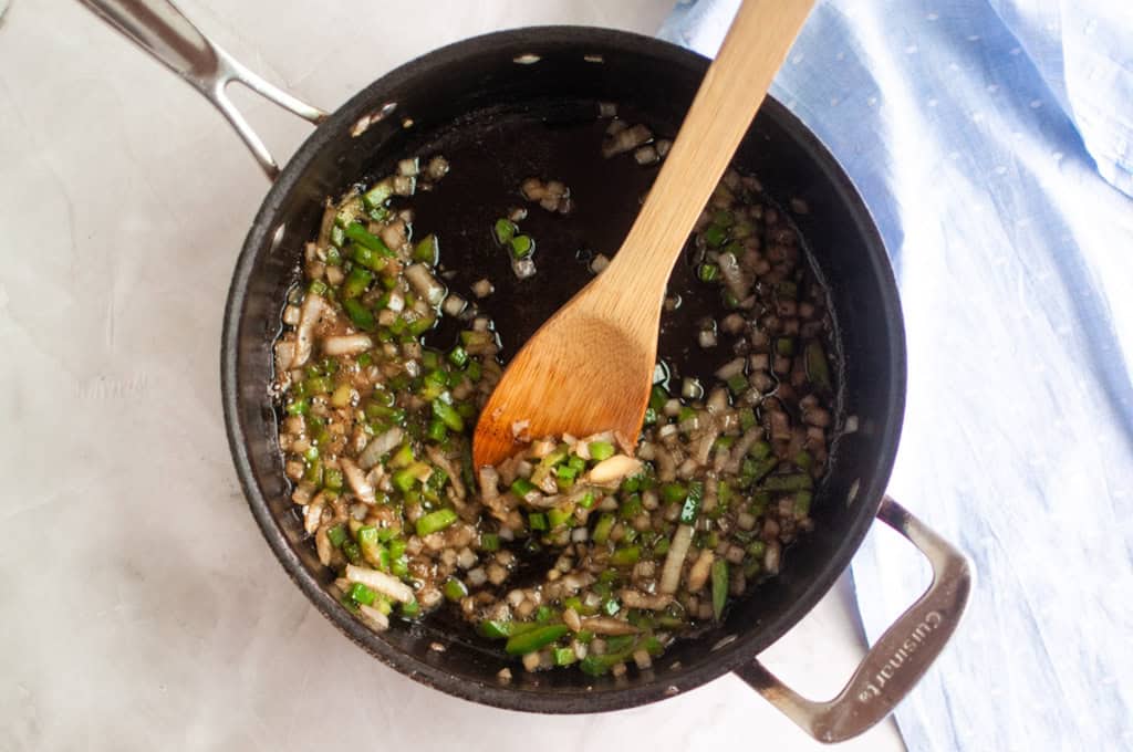 Chopped onions and green peppers are being sautéed in a black saucepan with a wooden spoon on a light surface.