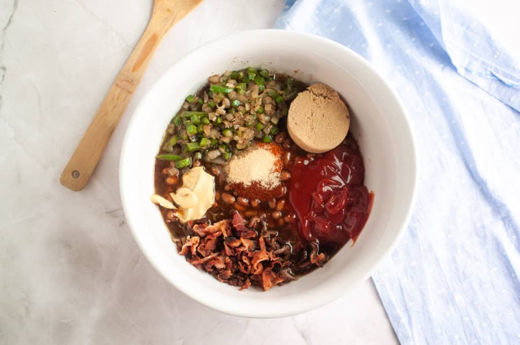 A white bowl contains chopped green peppers and onions, brown sugar, ketchup, cooked bacon pieces, seasonings, and beans, with a wooden spoon and blue cloth nearby.