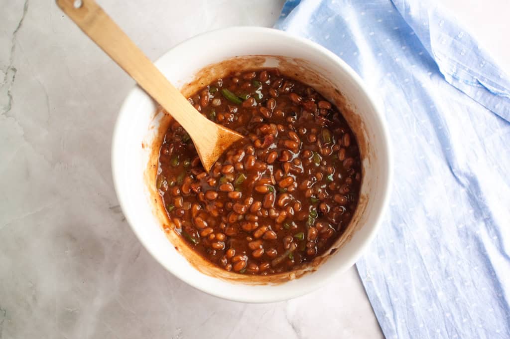 A white bowl filled with baked beans mixture and a wooden spoon sits on a marble countertop next to a light blue cloth.