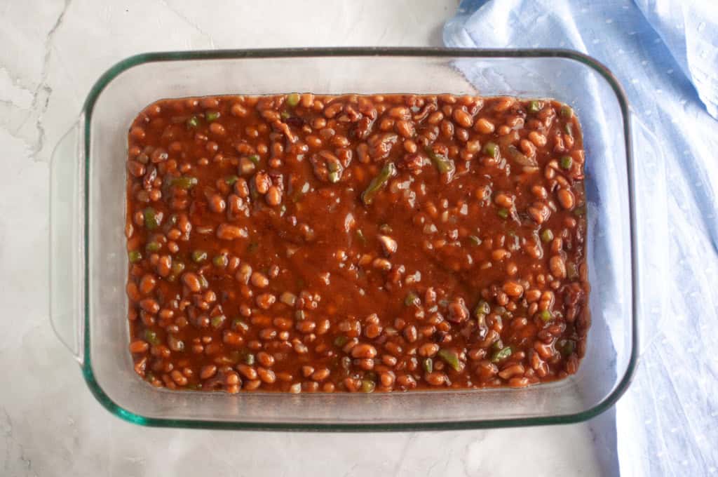 A glass baking dish filled with baked beans in sauce, placed on a light-colored surface next to a light blue cloth.