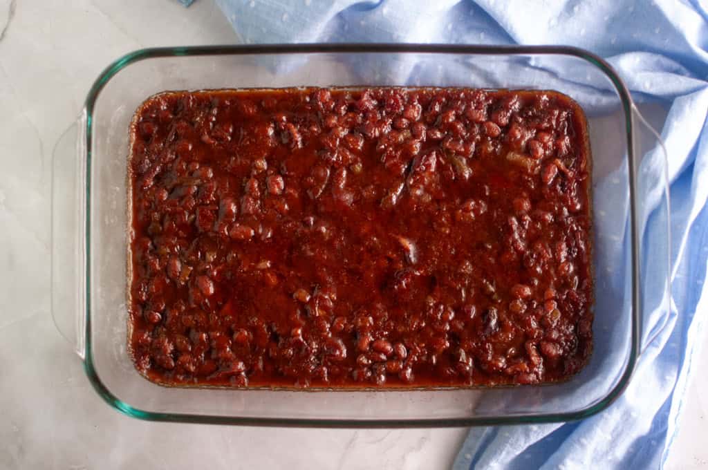A glass baking dish filled with baked beans in a thick, dark sauce, placed on a light-colored surface beside a blue cloth.