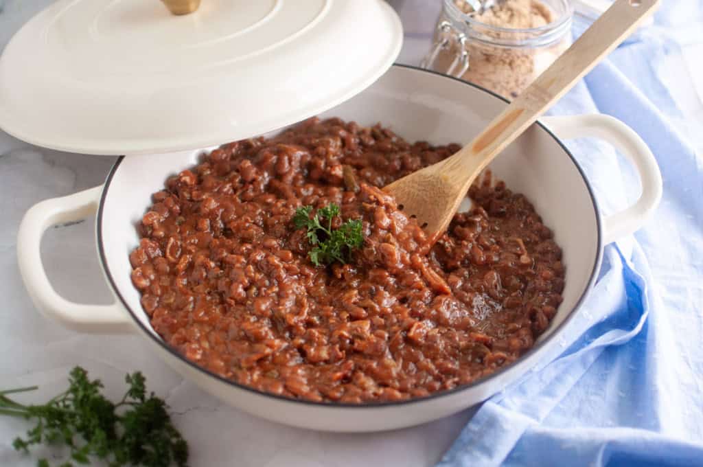 A white pot filled with cooked beans garnished with parsley, with a wooden spoon inside and a lid partially covering the pot.