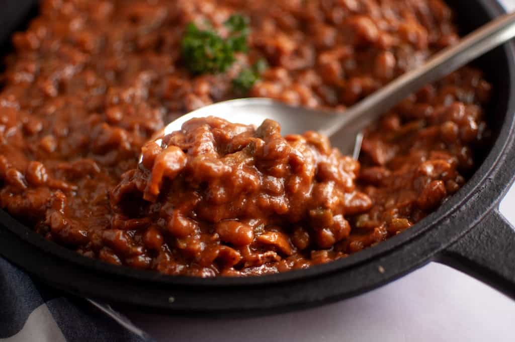 A close-up of a spoonful of baked beans in a black cast iron skillet, garnished with a small sprig of parsley.