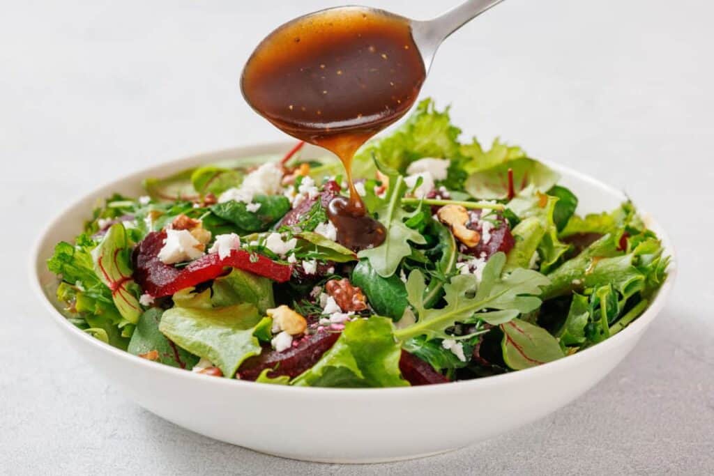 A spoon drizzles dark dressing over a salad in a white bowl containing mixed greens, beet slices, feta cheese, and walnuts.