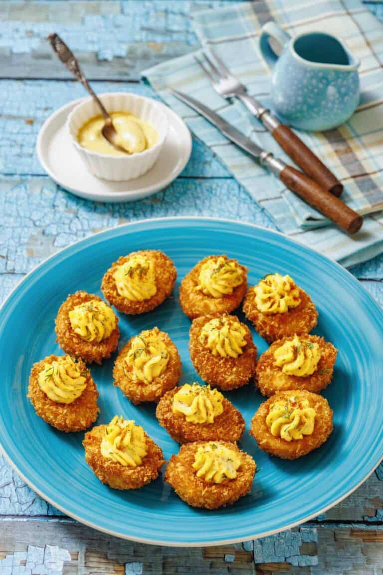 A blue plate with ten fried deviled eggs, garnished with herbs, is shown with a small bowl of mustard, forks, and a cloth.