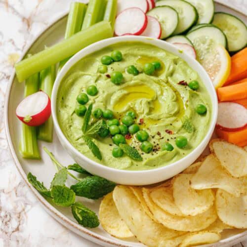 A bowl of Green Pea Hummus with Mint topped with peas, surrounded by sliced vegetables, potato chips, lemon slices, and fresh mint on a plate.