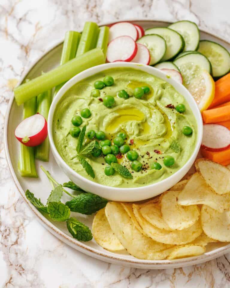 A bowl of Green Pea Hummus with Mint topped with peas, surrounded by sliced vegetables, potato chips, lemon slices, and fresh mint on a plate.