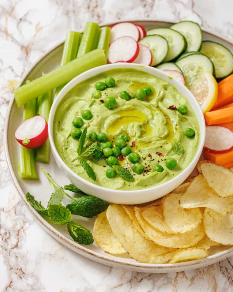 A bowl of Green Pea Hummus with Mint topped with peas, surrounded by sliced vegetables, potato chips, lemon slices, and fresh mint on a plate.
