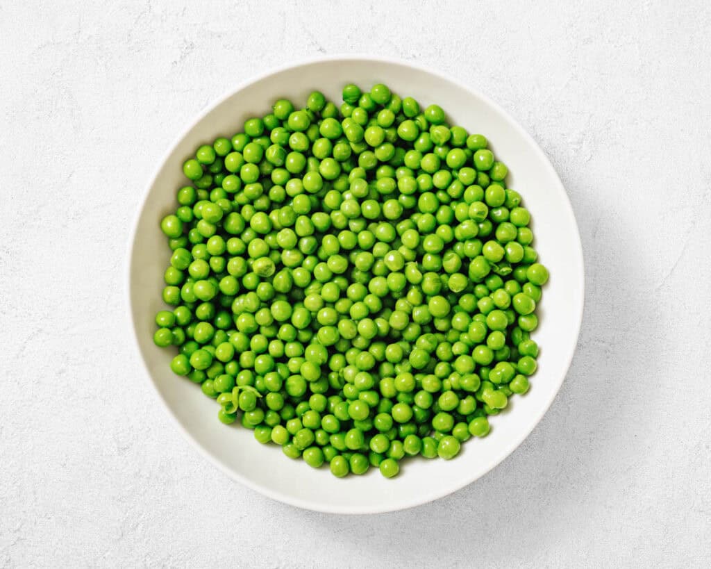 A white bowl filled with fresh green peas sits on a light textured surface, viewed from above.