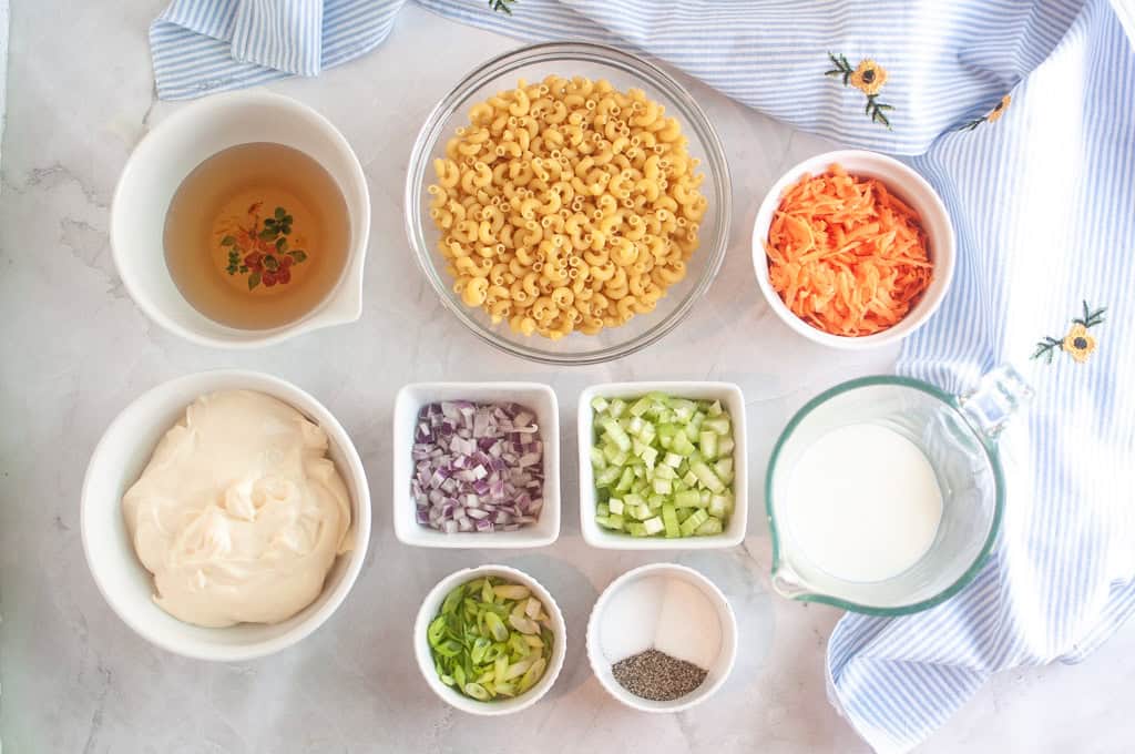 Overhead shot of ingredients for macaroni salad, including macaroni, carrots, mayonnaise, onions, celery, milk, seasonings, and broth, arranged on a light surface with a blue striped cloth.