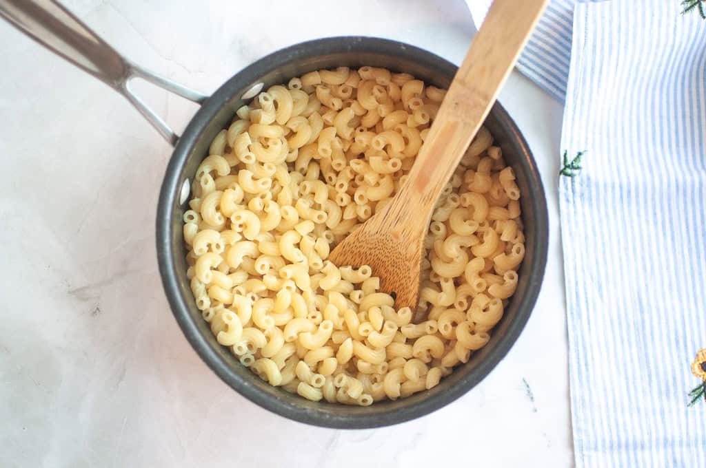 Overhead shot of cooked elbow macaroni in a dark pot with a wooden spoon, on a light surface with a striped cloth.