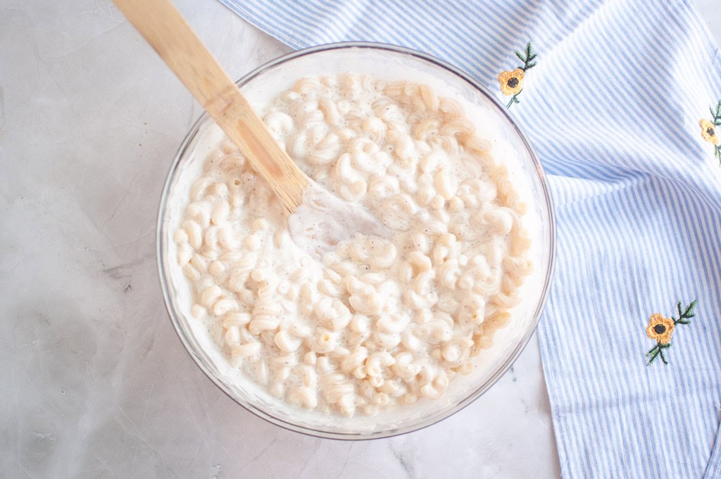 Overhead shot of macaroni salad being mixed in a glass bowl with a wooden spoon, with a striped cloth nearby.