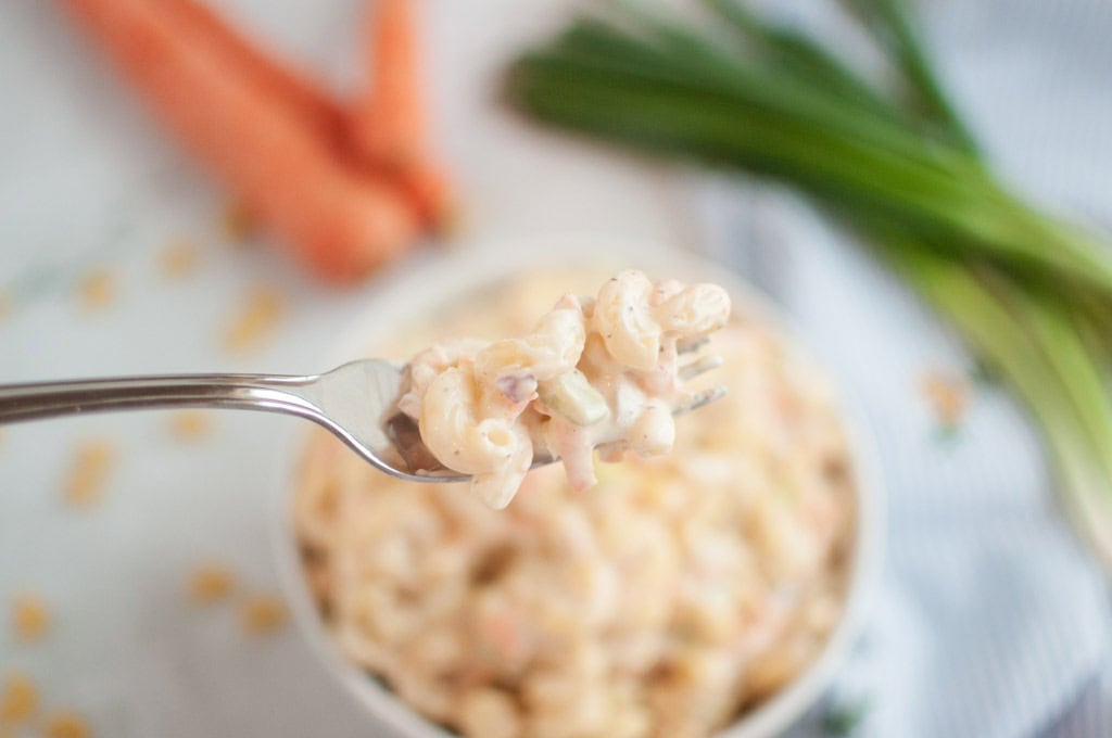 A close-up shot of a fork lifting a portion of macaroni salad, with a bowl of the salad and vegetables blurred in the background.