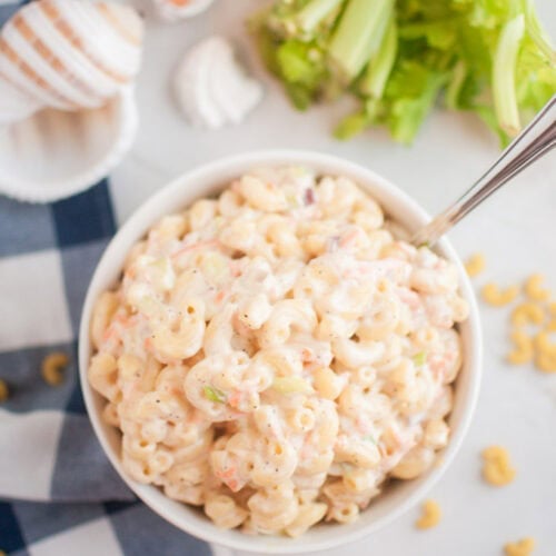 A white bowl filled with macaroni salad, containing elbow macaroni and a creamy dressing, is shown with a spoon inside. Celery and seashells are visible around the bowl.