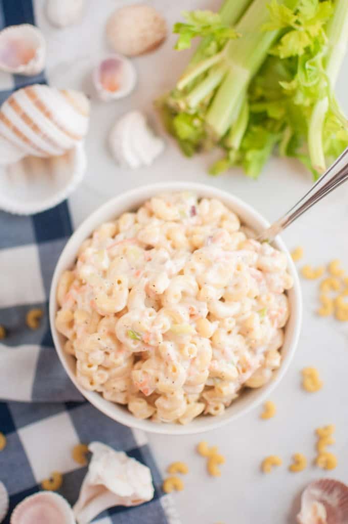 A white bowl filled with macaroni salad, containing elbow macaroni and a creamy dressing, is shown with a spoon inside. Celery and seashells are visible around the bowl.
