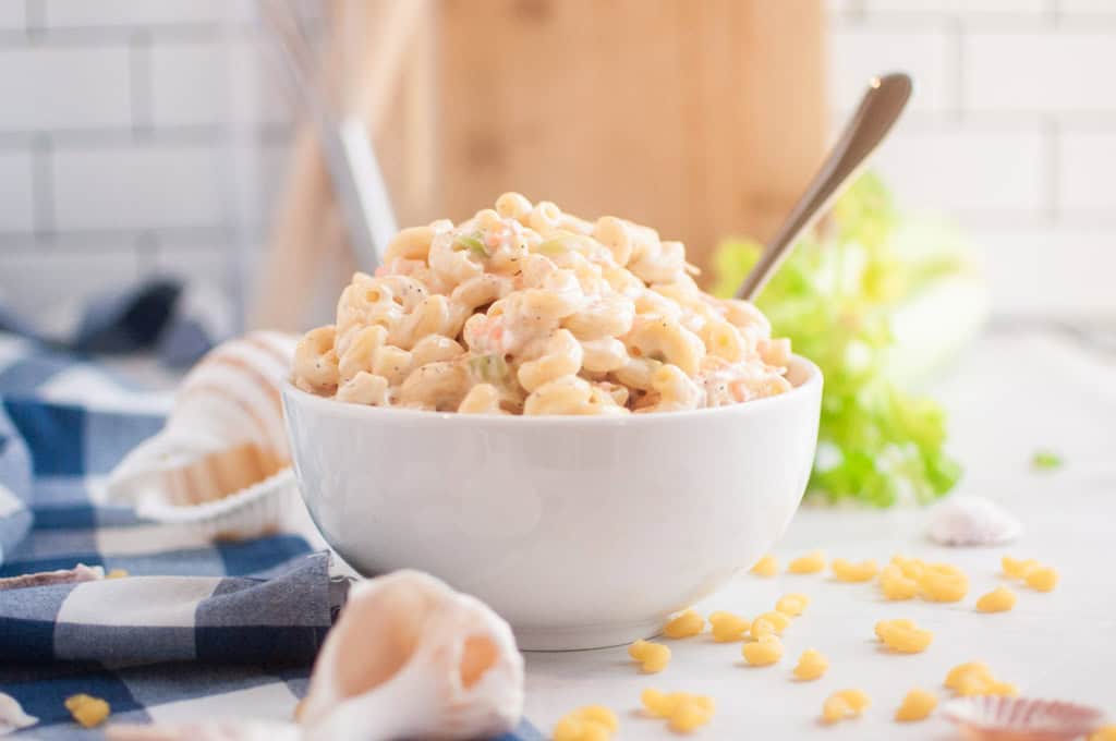 A white bowl filled with macaroni salad, containing elbow macaroni and a creamy dressing, is shown with a spoon inside. Garlic cloves and lettuce are visible around the bowl.