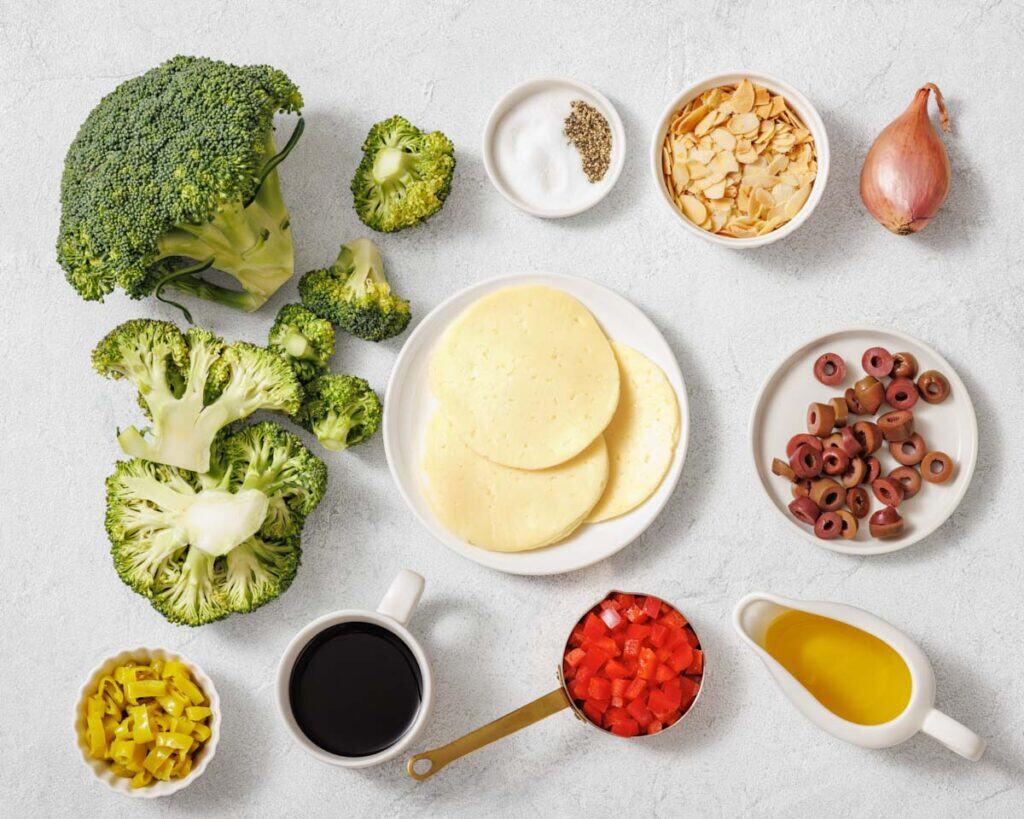 Various ingredients including broccoli, sliced cheese, olives, shallot, sliced almonds, chopped red peppers, olive oil, pepperoncini, balsamic vinegar, salt, and pepper arranged on a countertop.