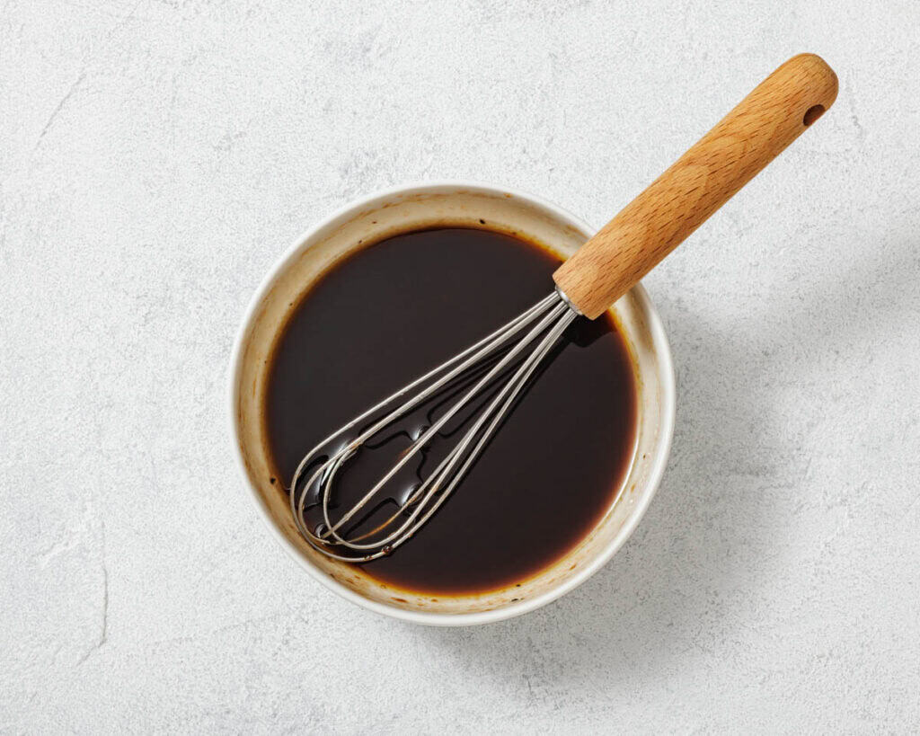 A metal whisk rests in a bowl filled with dark liquid, placed on a light textured surface.