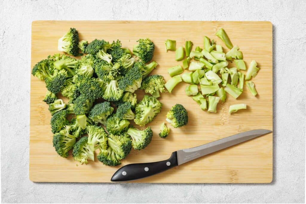 Chopped broccoli florets and stems on a wooden cutting board with a knife.