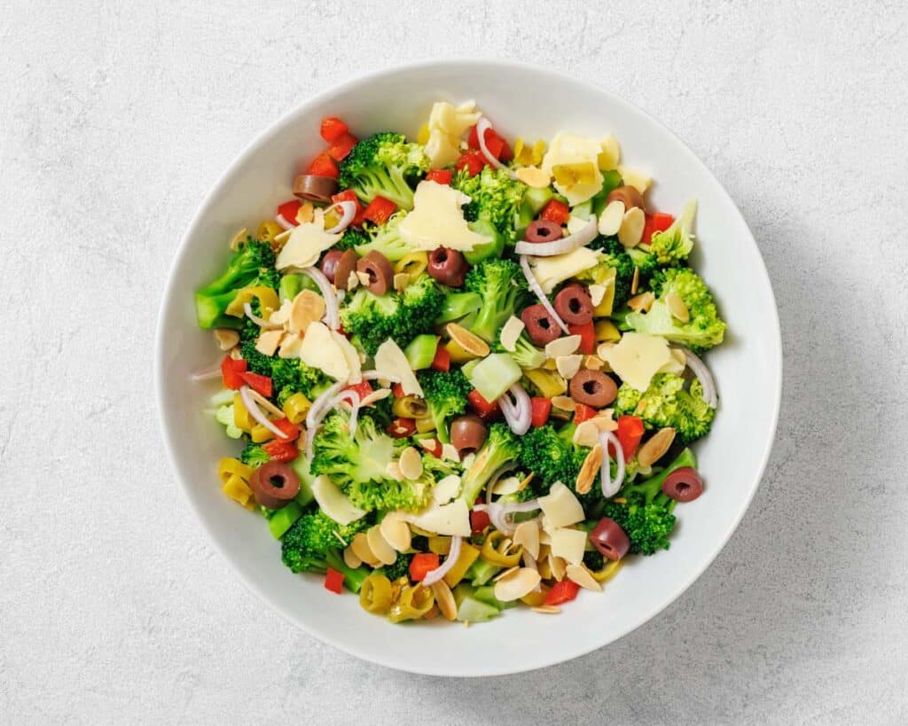 A bowl of broccoli salad with sliced olives, red bell peppers, onions, almonds, and shaved cheese on a light background.