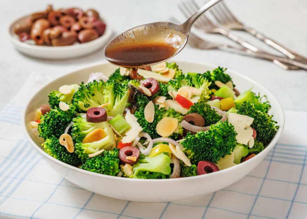 A bowl of broccoli salad with sliced olives, almonds, and vegetables is being drizzled with dressing from a spoon; a small dish of olives and forks are in the background.