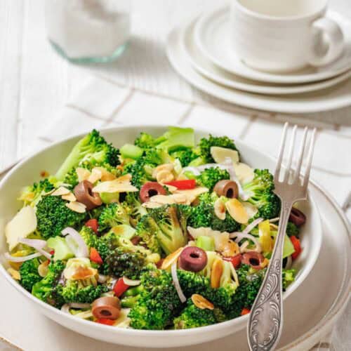 A bowl of broccoli salad with sliced almonds, olives, red onion, and red bell pepper sits on a plate with a fork, next to stacked dishes and a jar of salt.
