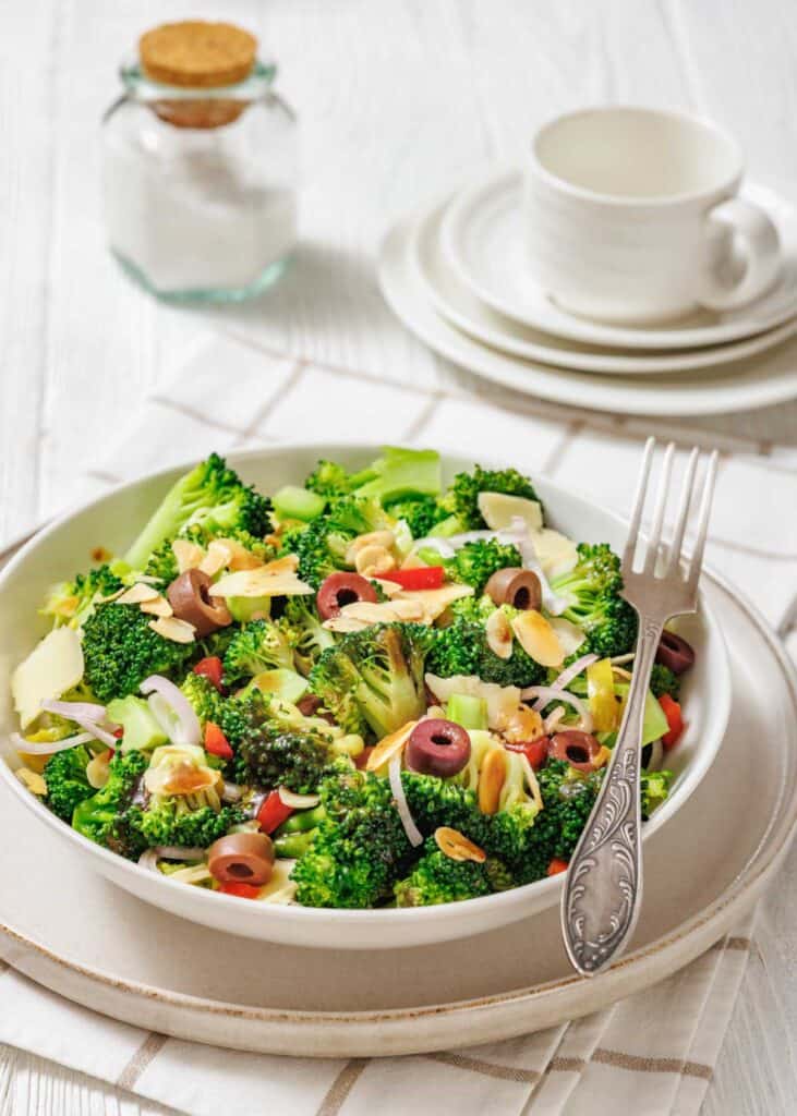 A bowl of broccoli salad with sliced almonds, olives, red onion, and red bell pepper sits on a plate with a fork, next to stacked dishes and a jar of salt.