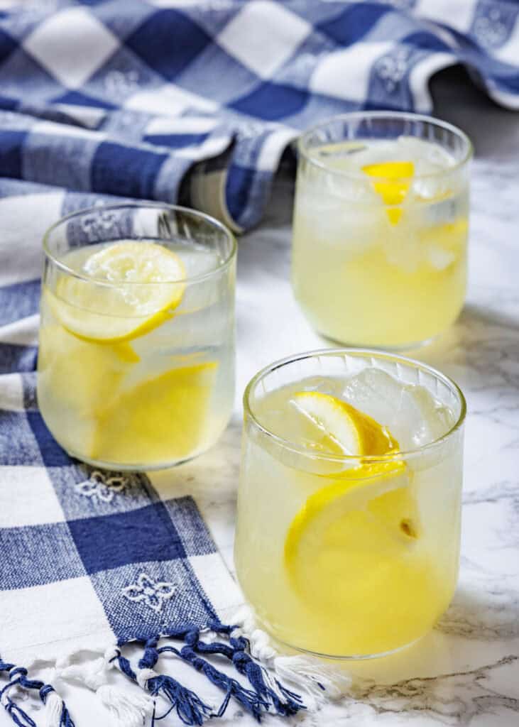Three short, clear glasses filled with lemonade and ice, garnished with lemon slices, are arranged on a blue and white checkered cloth.