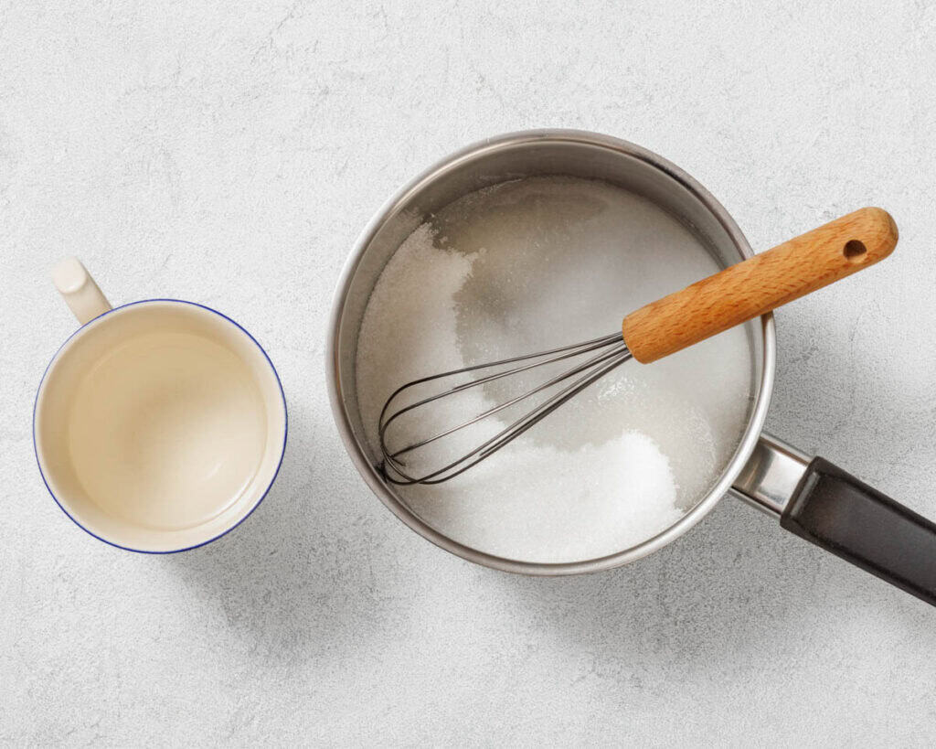 A top-down view of a metal saucepan with sugar and a whisk inside, next to a small cup of liquid, on a light gray textured surface.