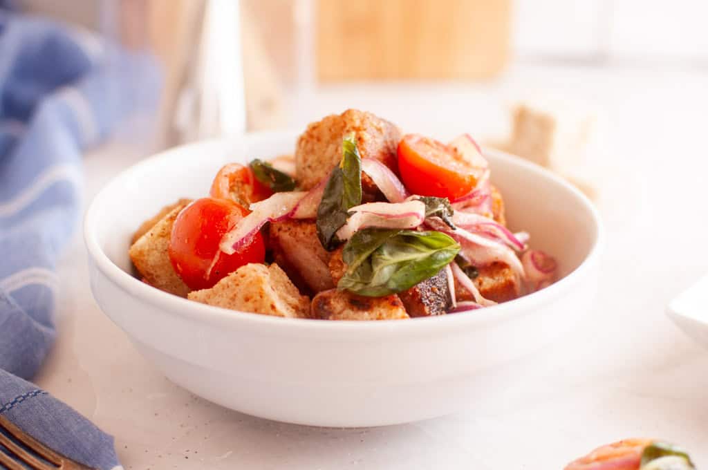 A white bowl filled with panzanella salad, featuring toasted bread cubes, cherry tomatoes, red onion, and basil leaves.