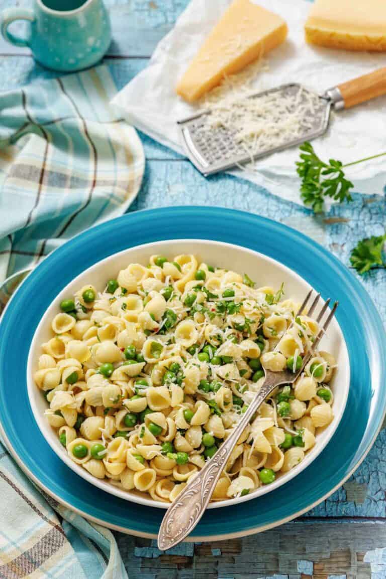 A bowl of pasta and peas with a fork, grater, and cheese on the side.