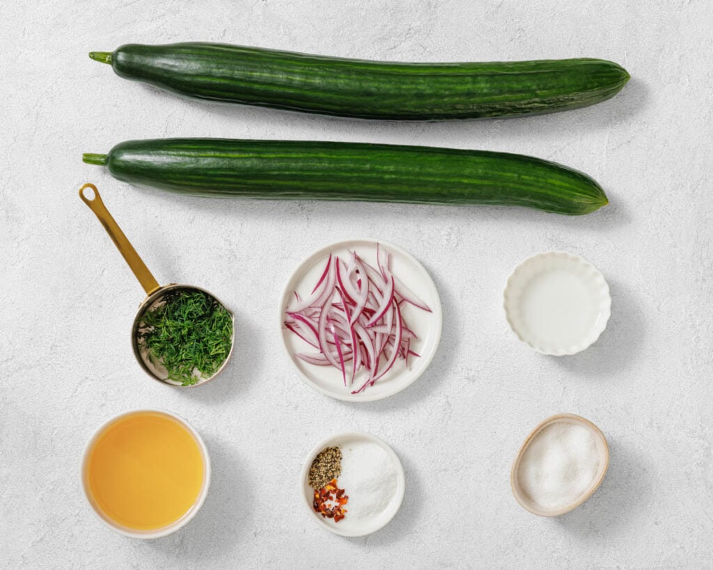 Two whole cucumbers, a plate of sliced red onions, chopped herbs, vinegar, sugar, salt, pepper, and spices are arranged on a white surface.