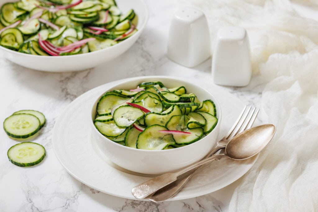 A bowl of Quick Marinated Cucumbers sits on a white plate with a fork and spoon beside it; salt and pepper shakers and another salad bowl are in the background.