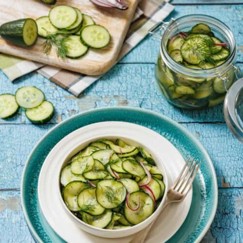 A bowl of Quick Marinated Cucumbers with red onion and dill sits on a plate with a fork, next to a cutting board with sliced cucumber and a jar of salad on a blue rustic table.