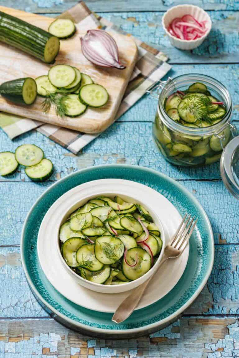 A bowl of Quick Marinated Cucumbers with red onion and dill sits on a plate with a fork, next to a cutting board with sliced cucumber and a jar of salad on a blue rustic table.