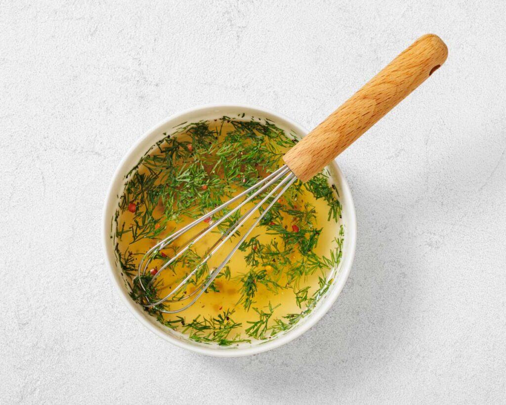 A white bowl containing a yellow liquid with chopped herbs, likely dill, and a metal whisk with a wooden handle resting inside.